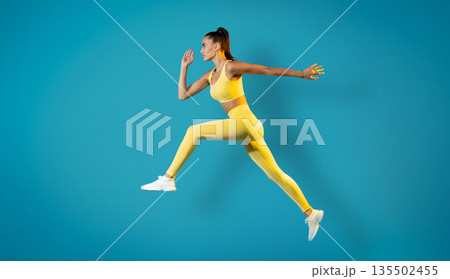 Athletic Woman Jumping Posing In Mid Air Training Over Blue Studio Background. Female Runner Leaping In Air. Workout And Sport Motivation Concept. Side View, Full Length 135502455