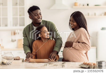 Happy Pregnant Black Woman And Her Husband And Daughter Baking Together Making Cookies And Rolling Out Dough In Modern Kitchen Indoor. Family Awaiting Baby Concept 135502476