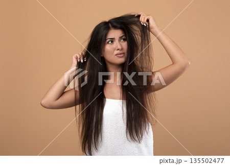 Frustrated armenian woman showing her damaged long locks on beige studio background. Young lady having bad hair day, upset over her messy hairdo. Hairdressing services concept 135502477
