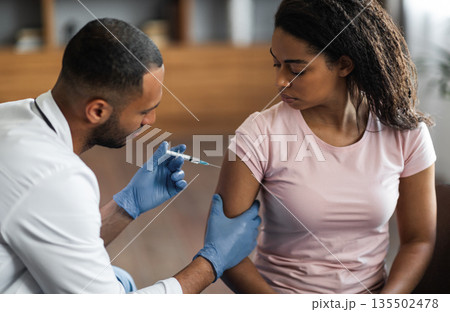 Handsome african american man making injection in shoulder for female patient, wearing blue gloves. Young black lady getting vaccinated against coronavirus at clinic, closeup shot 135502478