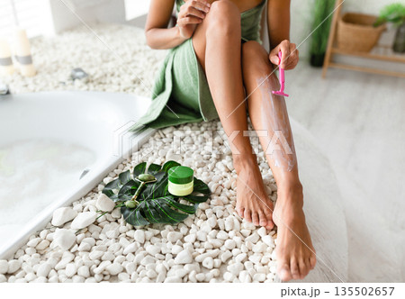 Unrecognizable young woman shaving legs with razor, removing unwanted hair, making epilation sitting on bathtub at home, closeup. Depilation and bodycare, domestic spa concept Unrecognizable young woman shaving legs with razor, removing unwanted hair, making epilation sitting on bathtub at home, closeup. Depilation and bodycare, domestic spa concept 135502657