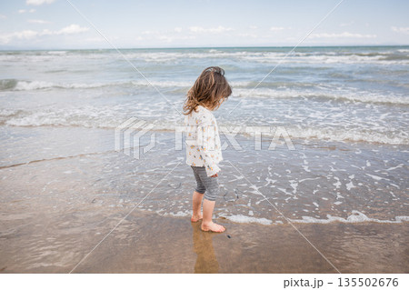 side view of little girl with brown hair standing on sand along the seashore side view of little girl with brown hair standing on sand along the seashore 135502676