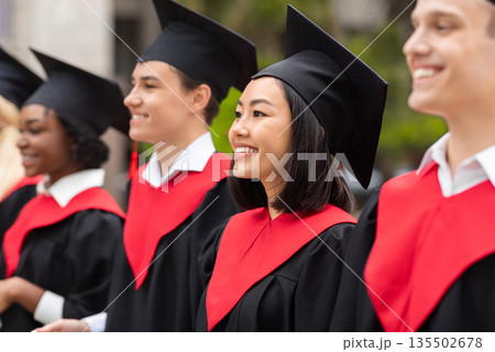 Multiracial group of cheerfully graduates having graduation ceremony, wearing black and red graduation robes and caps, standing in a row at university campus, closeup, panorama Multiracial group of cheerfully graduates having graduation ceremony, wearing black and red graduation robes and caps, standing in a row at university campus, closeup, panorama 135502678
