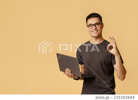 Excellent result, good feedback and win. Smiling young handsome male in glasses expresses emotions of success shows okay gesture and holds laptop, isolated on light background free space, studio shot 135502684