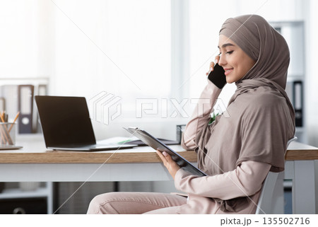 Muslim business lady in hijab holding clipboard, checking documentation and talking on mobile phone with partner while sitting at workplace in office Muslim business lady in hijab holding clipboard, checking documentation and talking on mobile phone with partner while sitting at workplace in office 135502716