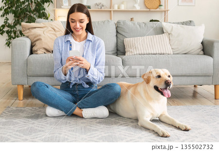 Lifestyle Concept. Portrait of smiling woman using smartphone sitting on the carpet with her purebred dog. Lady spending time at home, chatting at social media, texting sms or reading message Lifestyle Concept. Portrait of smiling woman using smartphone sitting on the carpet with her purebred dog. Lady spending time at home, chatting at social media, texting sms or reading message 135502732