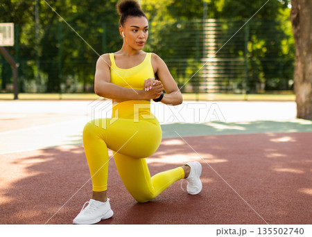 Sport And Healthy Lifestyle. Fit black woman doing curtsy lunges with twist, exercising outdoors on basketball stadium. Portrait of confident lady training in wireless earbuds, full body length 135502740