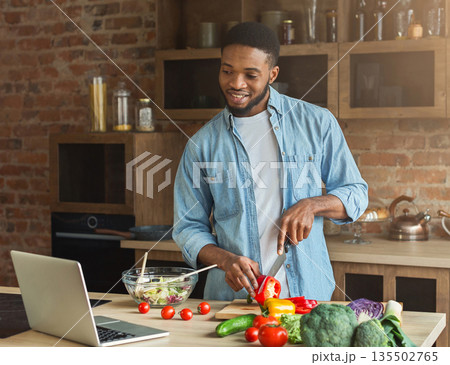 Happy african-american man preparing vegetable salad while looking on laptop 135502765