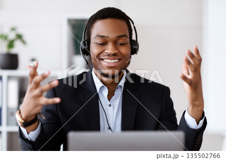 Closeup of inspired black businessman with headset having online conference, using laptop, talking to client or business partner, smiling at computer screen, business coach broadcasting from office Closeup of inspired black businessman with headset having online conference, using laptop, talking to client or business partner, smiling at computer screen, business coach broadcasting from office 135502766