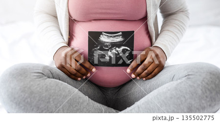 Unrecognizable black pregnant lady demonstrating her baby sonography photo while sitting on bed at home, showing firts photo of her child, enjoying happy maternity time, cropped image, closeup 135502775