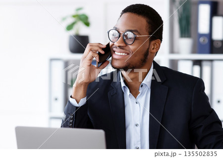 Cheerful african american young entrepreneur having business conversation on mobile phone, sitting in front of laptop in his office, taking notes and drinking coffee, copy space. Business lifestyle Cheerful african american young entrepreneur having business conversation on mobile phone, sitting in front of laptop in his office, taking notes and drinking coffee, copy space. Business lifestyle 135502835