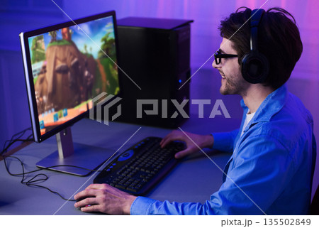 Male gamer playing shooter game on computer, wearing headphones and glasses Male gamer playing shooter game on computer, wearing headphones and glasses 135502849