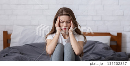 Unhappy millennial lady sitting on bed and massaging her temples, having headache or depression. Tired young woman suffering from insomnia, overwhelmed with negative thoughts Unhappy millennial lady sitting on bed and massaging her temples, having headache or depression. Tired young woman suffering from insomnia, overwhelmed with negative thoughts 135502887