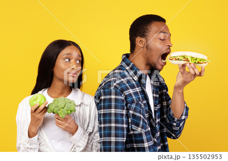 Healthy nutrition and fast food, diet and relationship. Funny millennial african american guy eats sandwich, wife with vegetables and fruits sadly looks at husband isolated on yellow background 135502953