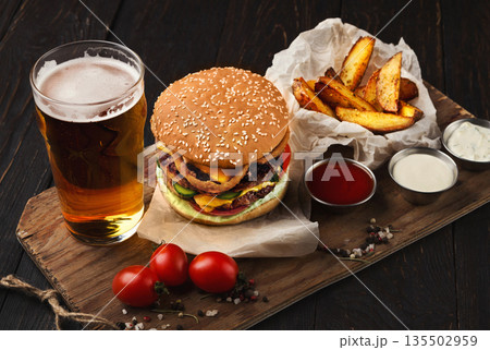 Fast food restaurant dish. Juicy burger, potato wedges, sauces and glass of cold beer on dark wooden background Fast food restaurant dish. Juicy burger, potato wedges, sauces and glass of cold beer on dark wooden background 135502959