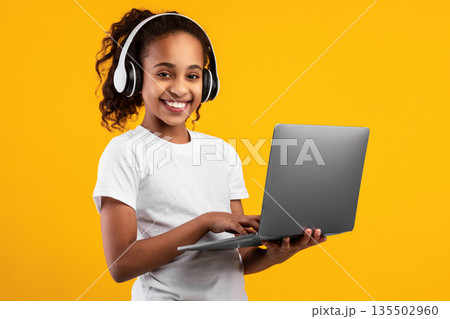 Kids And Technology Concept. Portrait of smiling black girl holding and using laptop computer, wearing wireless headphones, studying, watching online course, isolated over yellow studio wall Kids And Technology Concept. Portrait of smiling black girl holding and using laptop computer, wearing wireless headphones, studying, watching online course, isolated over yellow studio wall 135502960