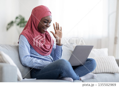 Cheerful black muslim lady in hijab making video call on laptop at home, waving at screen, chatting with family or friends, enjoying online communication, sitting on couch in living room, copy space 135502969
