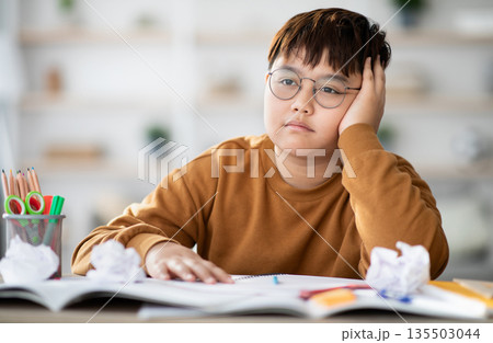 Cute overweight japanese boy teenager doing school project at home, sitting at desk with books and notebooks, got tired and bored, looking for creative solutions, closeup photo, copy space 135503044
