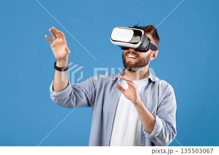 Happy bearded young man using VR headset to explore augmented reality on blue studio background. Cheerful millennial guy playing virtual online game, having fun with modern technologies 135503067