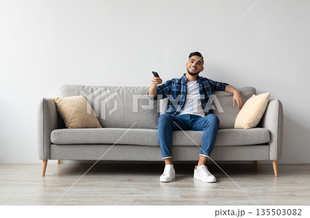 Portrait of Arab guy watching TV show or film, holding remote control, switching channels. Young smiling man spending weekend free time sitting on comfortable sofa at home in living room, copy space Portrait of Arab guy watching TV show or film, holding remote control, switching channels. Young smiling man spending weekend free time sitting on comfortable sofa at home in living room, copy space 135503082