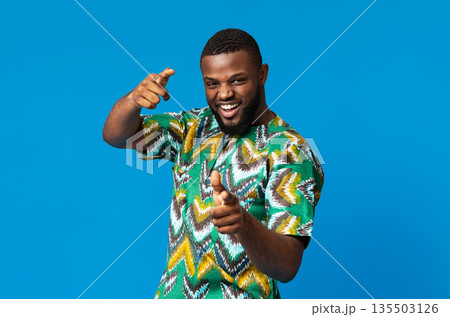 Playful black young man in colorful african costume, indicating to camera on blue studio background, empty space. Happy african american charismatic guy demonstrating traditional bright clothes 135503126