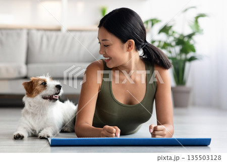 Beautiful puppy exercising with its owner athletic asian woman, cheerful young lady in sportswear laying on yoga mat in living room and looking at cute jack russel terrier laying by, copy space Beautiful puppy exercising with its owner athletic asian woman, cheerful young lady in sportswear laying on yoga mat in living room and looking at cute jack russel terrier laying by, copy space 135503128
