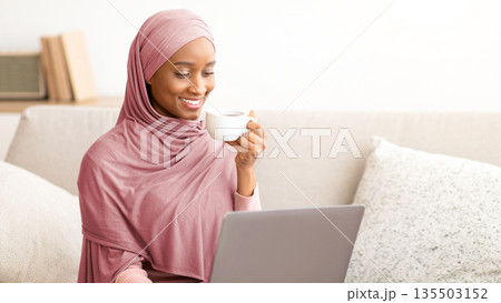 Online life concept. Black woman in hijab sitting on couch with laptop, drinking coffee, working or studying from home. Attractive African American lady in headscarf having business meeting on web 135503152