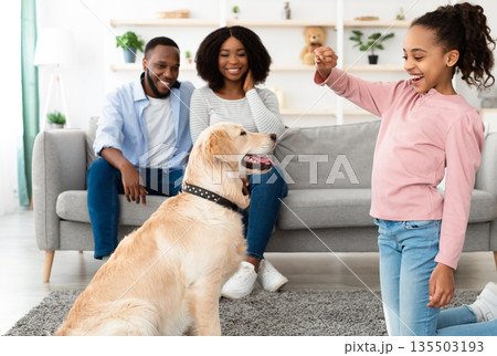 Dog Training Commands. Happy black girl teaching pet at home in living room, playing with golden retriever and rewarding him with treats, standing indoors giving animal food, parents sitting on sofa Dog Training Commands. Happy black girl teaching pet at home in living room, playing with golden retriever and rewarding him with treats, standing indoors giving animal food, parents sitting on sofa 135503193