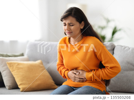 Concerned unhappy young indian woman in casual suffering from stomach-ache, sitting on sofa at home. Female holding tummy, undergoing belly pain and discomfort, suffer from menstruation, copy space Concerned unhappy young indian woman in casual suffering from stomach-ache, sitting on sofa at home. Female holding tummy, undergoing belly pain and discomfort, suffer from menstruation, copy space 135503208