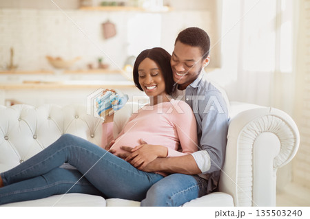Loving black man hugging his pregnant wife with baby booties on sofa at home. Romantic African American couple with cute tiny shoes cuddling together on couch, enjoying peaceful moments as family Loving black man hugging his pregnant wife with baby booties on sofa at home. Romantic African American couple with cute tiny shoes cuddling together on couch, enjoying peaceful moments as family 135503240