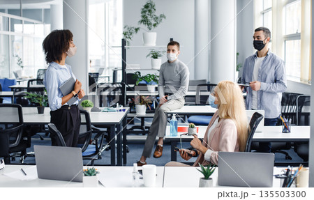 Report on work, meeting or team building during covid-19 outbreak and social distancing. Young african american lady with tablet speaks to colleagues in modern office interior with gadgets, panorama 135503300