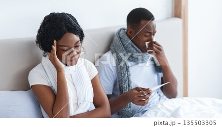 Sick african american couple wrapped in scarfs sitting in bed at bedroom, measuring fever and sneezing noses. Young black family of two got sick, staying at home, seasonal viruses and flu concept 135503305