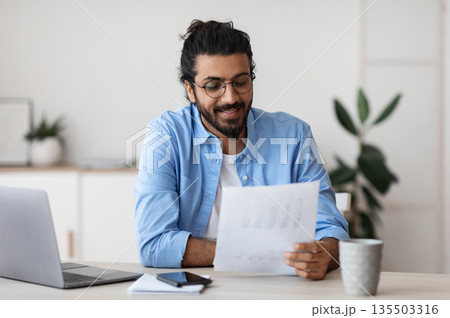 Young Arab Male Self-Entrepreneur Working With Papers At Desk In Home Office, Checking Annual Financial Reports. Eastern Millennial Man Reading Documents, Sitting At Table With Computer, Copy Space Young Arab Male Self-Entrepreneur Working With Papers At Desk In Home Office, Checking Annual Financial Reports. Eastern Millennial Man Reading Documents, Sitting At Table With Computer, Copy Space 135503316