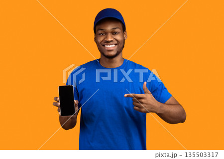 Recommending Service. Smiling african american delivery guy holding smartphone with blank screen for mockup, wearing blue cap and uniform, pointing at black empty monitor isolated on orange background Recommending Service. Smiling african american delivery guy holding smartphone with blank screen for mockup, wearing blue cap and uniform, pointing at black empty monitor isolated on orange background 135503317