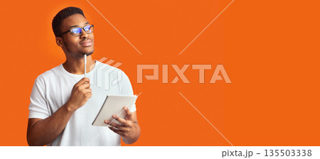 Pensive Redactor. Dreaming afro man in glasses taking notes, writing ideas, looking at copyspace, orange wall, panorama 135503338