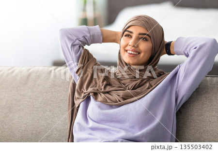 Rest Concept. Muslim lady wearing headscarf sitting on comfortable couch at home in living room. Happy islamic woman relaxing on sofa, leaning back, enjoying weekend free time or break from work Rest Concept. Muslim lady wearing headscarf sitting on comfortable couch at home in living room. Happy islamic woman relaxing on sofa, leaning back, enjoying weekend free time or break from work 135503402