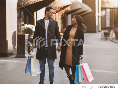 Great Black Friday shopping for seasonal sales. Happy african american husband and wife hold hands each other and colored purchases bags walking on street near city mall, outdoor, copy space 135503403