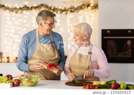 Christmas Celebration. Senior Husband Giving Gift To Wife Surprising Her On Xmas Eve Celebrating Holiday Together In Kitchen At Home. Elderly Spouses Congratulating Each Other On New Year Concept Christmas Celebration. Senior Husband Giving Gift To Wife Surprising Her On Xmas Eve Celebrating Holiday Together In Kitchen At Home. Elderly Spouses Congratulating Each Other On New Year Concept 135503417