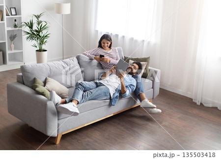 High angle view of relaxed cheerful stylish millennial eastern lovers man and woman chilling on couch in cozy living room, using gadgets at home, scrolling on social media, reading online, copy space 135503418