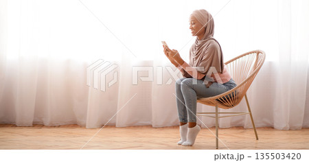 Millennial black lady in hijab using cellphone at home, working or studying remotely, free space. African American woman in Muslim headscarf checking email, sending message, having online video call 135503420