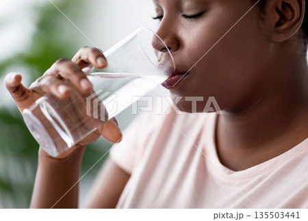Keep hydrated for healthier living. Overweight black woman drinking clear mineral water from glass indoors. Plus size African American lady taking care of her wellbeing at home 135503441