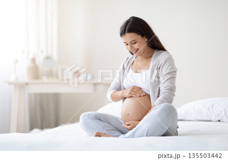 Happy pregnant woman sitting on bed in bedroom at home, hugging her big tummy. Happiness and feeling joyful as emotional well affect to baby neurological and psychological development, empty space 135503442