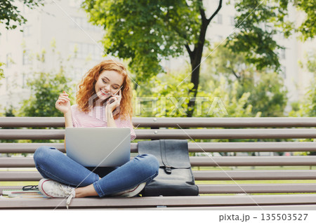 Young woman sitting on bench, talking on smartphone, working on laptop outdoors. Technology, communication, education and remote working concept, copy space Young woman sitting on bench, talking on smartphone, working on laptop outdoors. Technology, communication, education and remote working concept, copy space 135503527