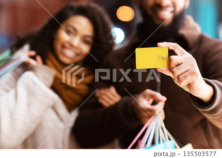 Easy Shopping. Portrait of smiling black couple showing debit credit card and holding shopper bags, walking outside in the city near mall in the evening, selective focus, blurred background Easy Shopping. Portrait of smiling black couple showing debit credit card and holding shopper bags, walking outside in the city near mall in the evening, selective focus, blurred background 135503577