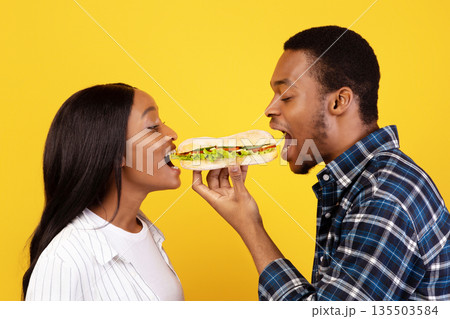 Romantic date and fast food. Cute millennial african american man and woman eat one sandwich for two at covid-19 quarantine, isolation on yellow background, studio shot, profile, copy space 135503584
