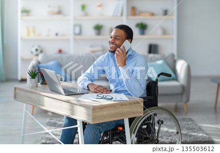 Joyful disabled black guy in wheelchair having phone conversation while working online on laptop from home. Young handicapped businessman talking with client on mobile phone Joyful disabled black guy in wheelchair having phone conversation while working online on laptop from home. Young handicapped businessman talking with client on mobile phone 135503612