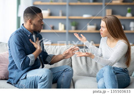 Interracial Couples Problems. Millennial Black Man And White Woman Arguing At Home, Emotional Multicultural Lovers Quarreling While Sitting On Couch In Living Room, Suffering Relationship Crisis 135503633