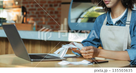 Financial difficulties and problems of small businesses during covid-19 lockdown. Millennial african american woman owner manager in apron works with account and bookkeeping in cafe, panorama, cropped 135503635