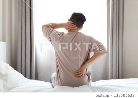 Tired man suffering from back pain after sleeping in bedroom, waking up in morning, sitting on bed and touching lower back and neck, experiencing body aches, wearing pajamas, copy space, back view 135503638