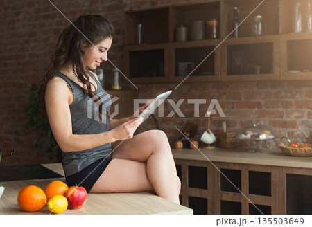 Happy young woman eating fruits and using digital tablet in loft kitchen. Early morning, breakfast, routine concept 135503649